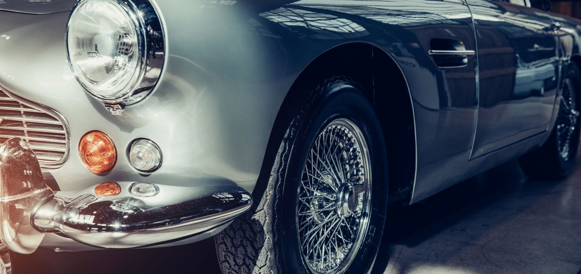 Close-up of a classic silver sports car showing details of the shiny chrome grille, headlights, and spoked wheel in a softly illuminated garage.