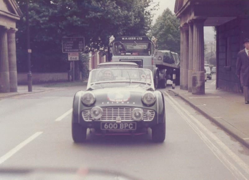 A vintage car drives on a city street with other vehicles and a pedestrian under an overpass. the scene captures a nostalgic, mid-20th-century urban setting.