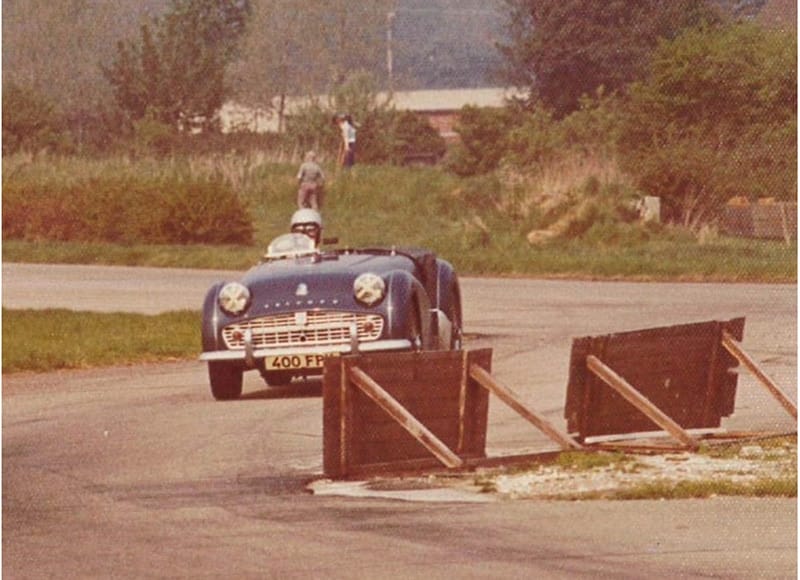 Vintage sports car driving on a track with a person standing in the background, near rustic wooden barriers and greenery.