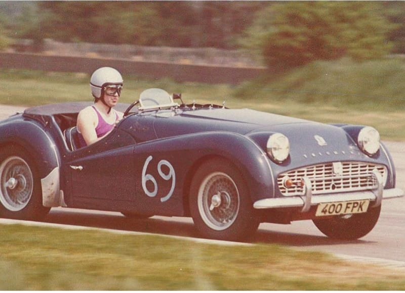 A vintage photograph of a smiling man wearing a helmet and sunglasses, driving a classic number 69 sports car with the license plate "400 fpk" on a racetrack.