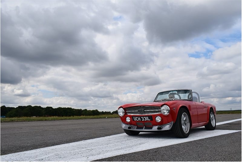 A classic red convertible car parked on an airstrip under a cloudy sky.
