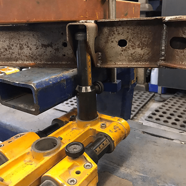 A close-up shot of a yellow hydraulic tool being used to bend a rusted metal beam. The tool is positioned underneath the beam, and a gauge indicates the angle of the bend. The setup is in a workshop environment with various machinery and tools in the background.