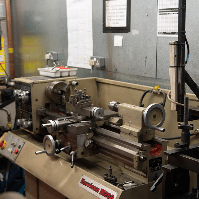 A metal lathe machine with various controls and attachments in a workshop setting. The machine is labeled "Harrison M250". Surrounding the lathe are tools and equipment, with a gray metal wall in the background that contains posted charts and notices.