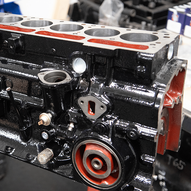 The image displays a close-up view of a metal engine block with various exposed components and cavities. The engine block is painted black with some red accents inside the cylindrical chambers, and there are visible bolt holes and connection points on its surface.